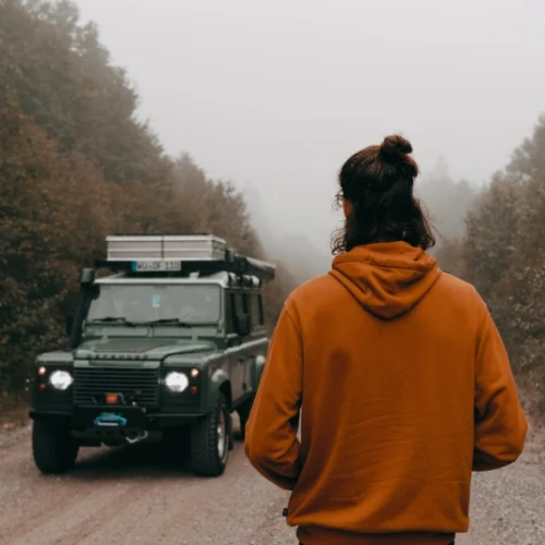 Man with orange hoodie standing with back, looking at Land Rover Defender. Mystic scenery around with fog all around.