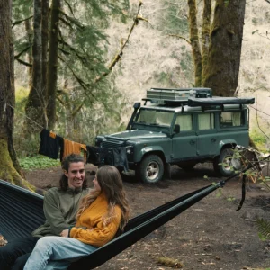 Bianca and Tim sitting in a hammock in a green forest. Their green Land Rover Defender sits in the background. They're surrounded by trees and a few clothes hanging on a rope to dry.