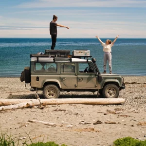 The green Land Rover Defender parked on a beach in the centre of the picture. The ocean is in the background. Tim stands on the roof of the car pointing towards Bianca who's standing on the hood with arms raised in the air filled with happiness.