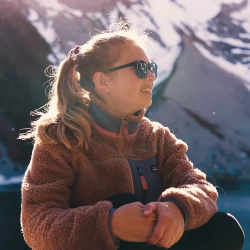 Brand shoot sun glasses: Bianca wearing sun glasses at a mountain lake with snowy mountains in the back. The sun rays light up her face and hair.