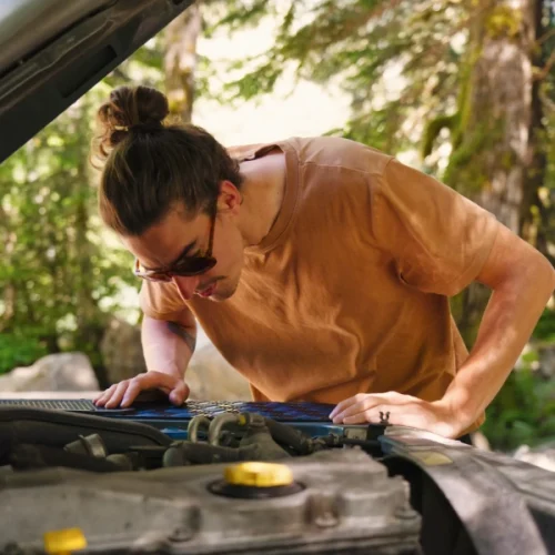 Brand shoot sun glasses: Tim checks under the Hood of our Land Rover Defender. He wears a brown Shirt and sun glasses. Trees in the back.