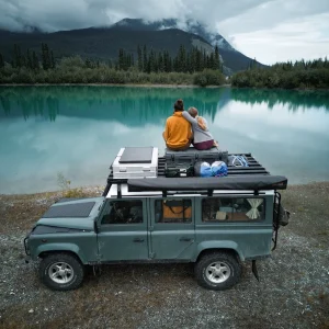 Bianca and Tim sitting on the roof of their Land Rover Defender starring in the distance towards a tall Mountain. Between them and the Mountain is a turquoise blue lake. The image was taken in the Yukon Territory, Canada.