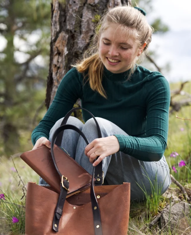 portfolio main Brand shoot leather goods: Bianca sits next to a tree. She holds a brown leather bag, takes out a leather journal and smiles.