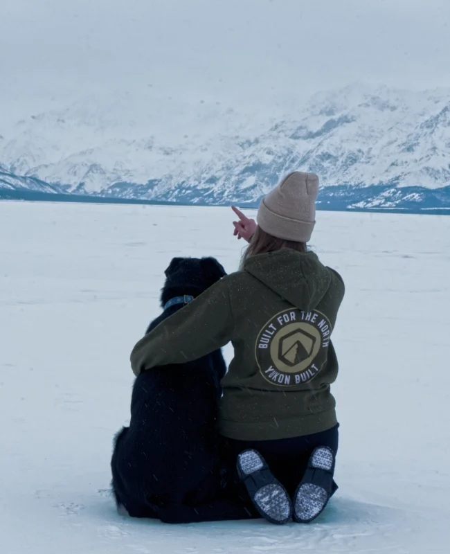 A woman in a green hoody, wearing a toque is sitting with her dog on a frozen lake in northern Canada. Mountains covered in snow fill the background of the image