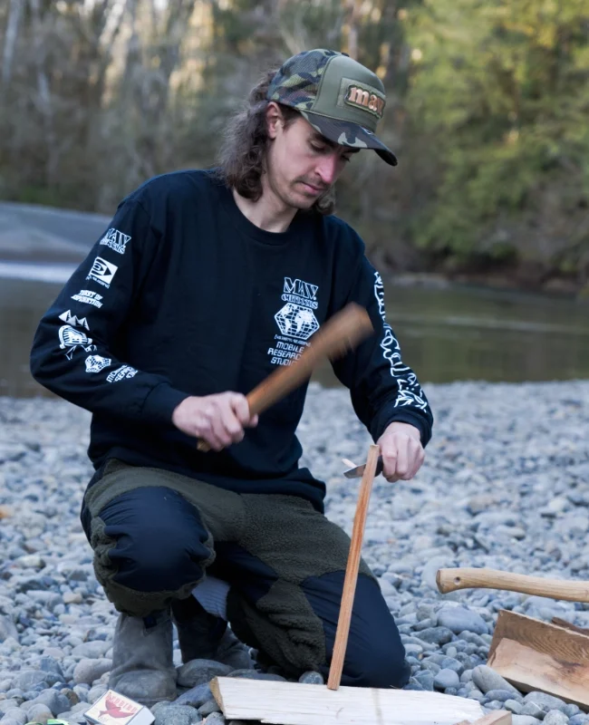 Brand shoot apparel: Tim is preparing a bonfire, he splits a small piece of wood in two with his knife. He wears a camo cap and a black long sleeve. The backdrop is a riverbed with gravel and trees.